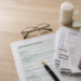 German tax forms and receipts on a wooden desk, with a pen, eyeglasses, a candle, and a white lily nearby.