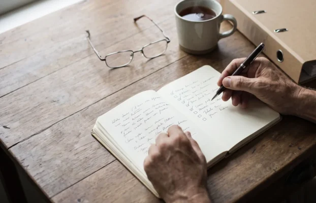 Hände mit Stift über einem Notizbuch mit Checkliste, daneben Lesebrille, eine Tasse Tee und ein Aktenordner auf einem Holztisch