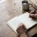 Hände mit Stift über einem Notizbuch mit Checkliste, daneben Lesebrille, eine Tasse Tee und ein Aktenordner auf einem Holztisch