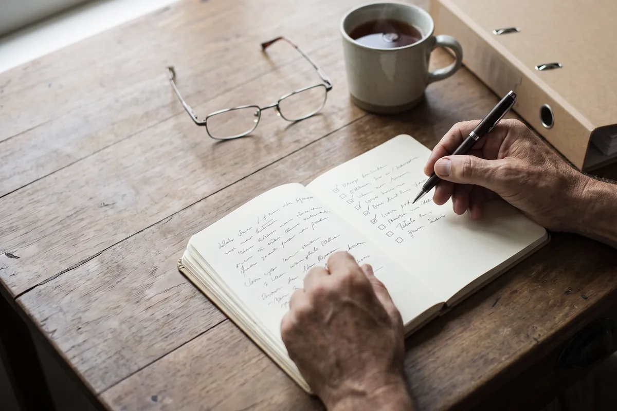 Hände mit Stift über einem Notizbuch mit Checkliste, daneben Lesebrille, eine Tasse Tee und ein Aktenordner auf einem Holztisch